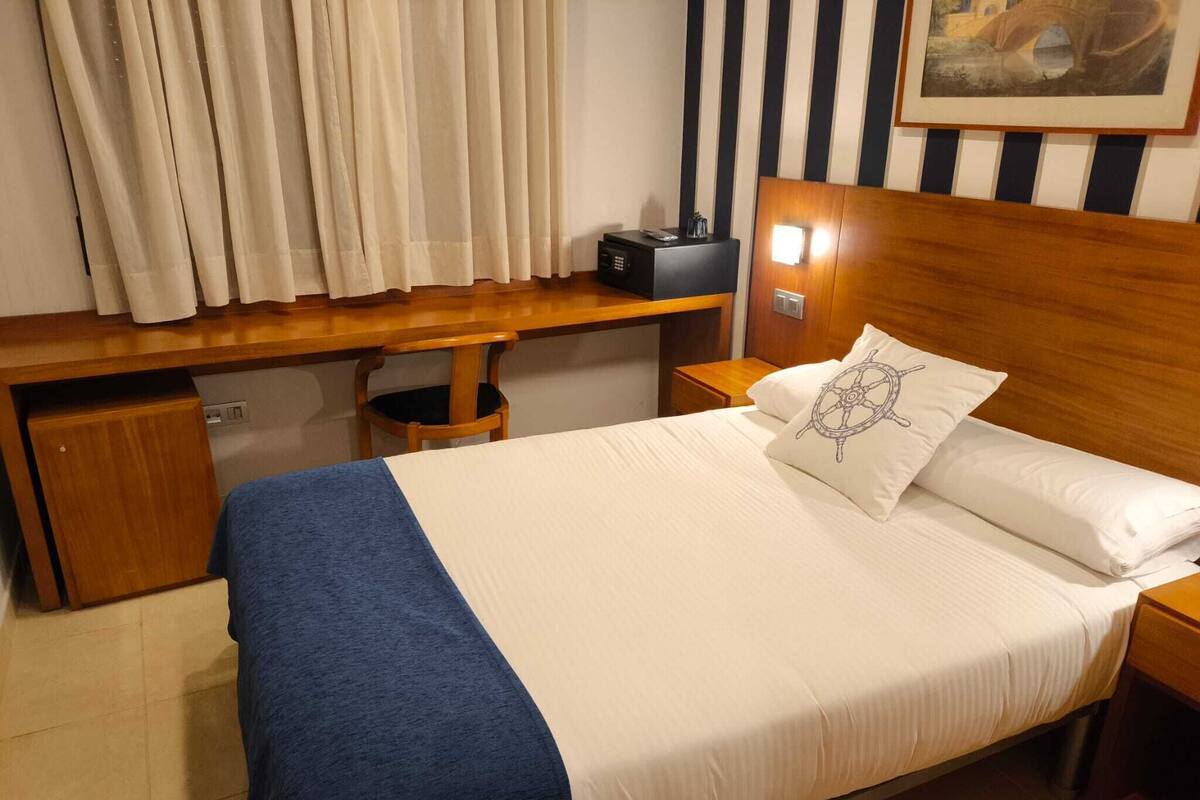 A neatly made bed with a blue blanket, a wooden nightstand with a radio, and a painting on the wall.