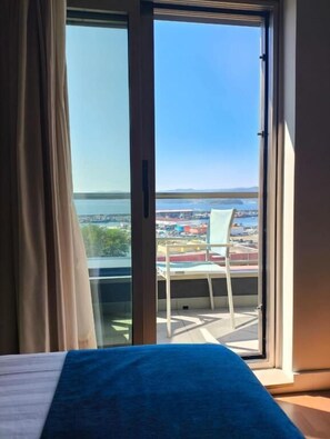 A room with a bed, a view of a cityscape through a large window, and a balcony with a chair.