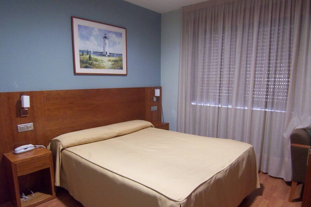 A hotel room with a bed, a nightstand, a lamp, a painting of a lighthouse, and a window with curtains.