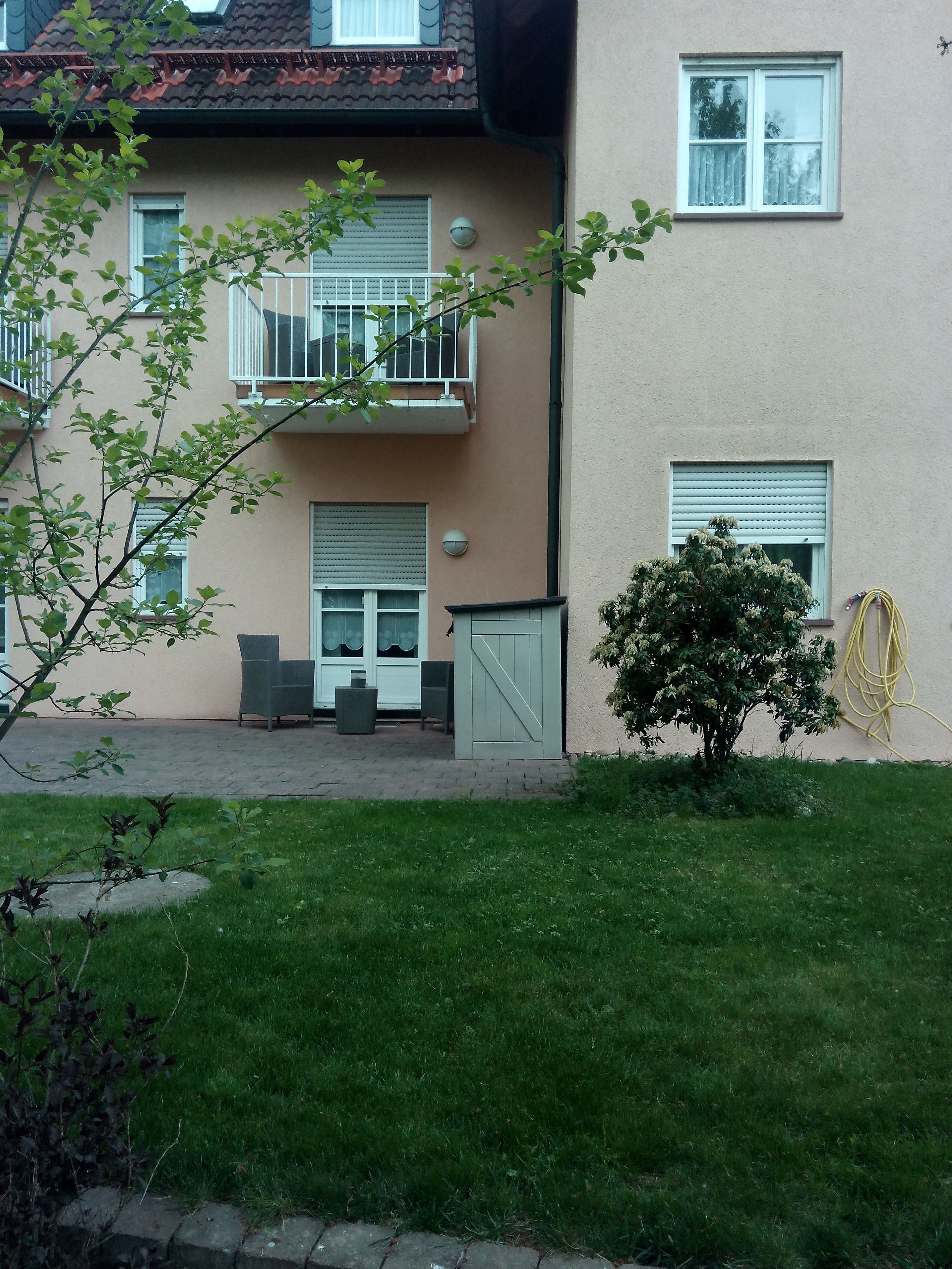 A residential building with a balcony, a small garden, and a hose on the wall.