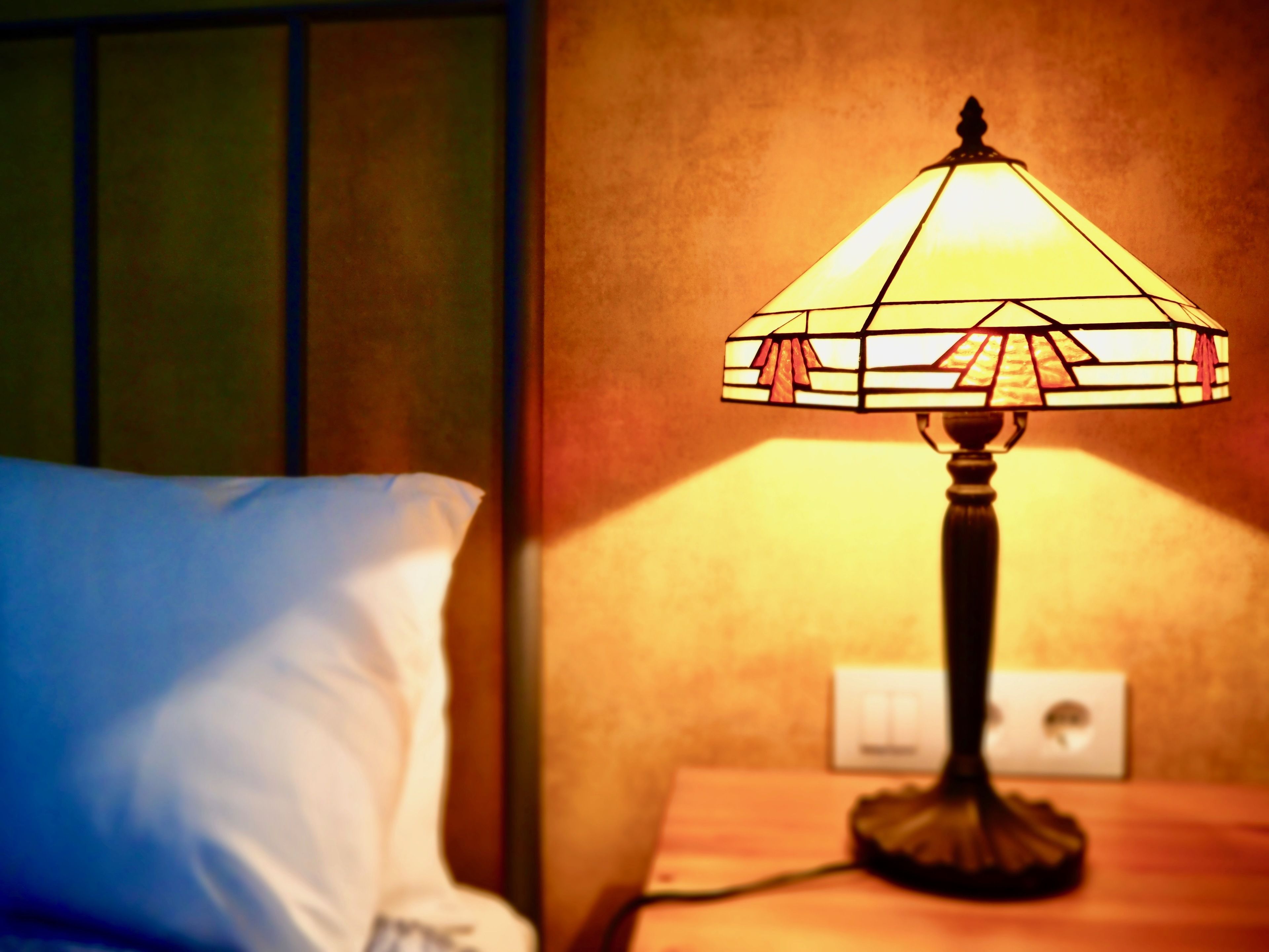 A bedside lamp with a stained glass shade on a wooden nightstand next to a bed with white pillows.