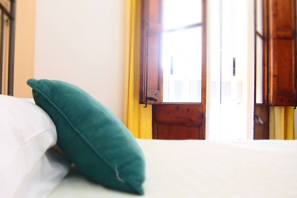 Una cama con sábana blanca y almohada azul turquesa, una puerta de madera y una ventana con cortinas amarillas.