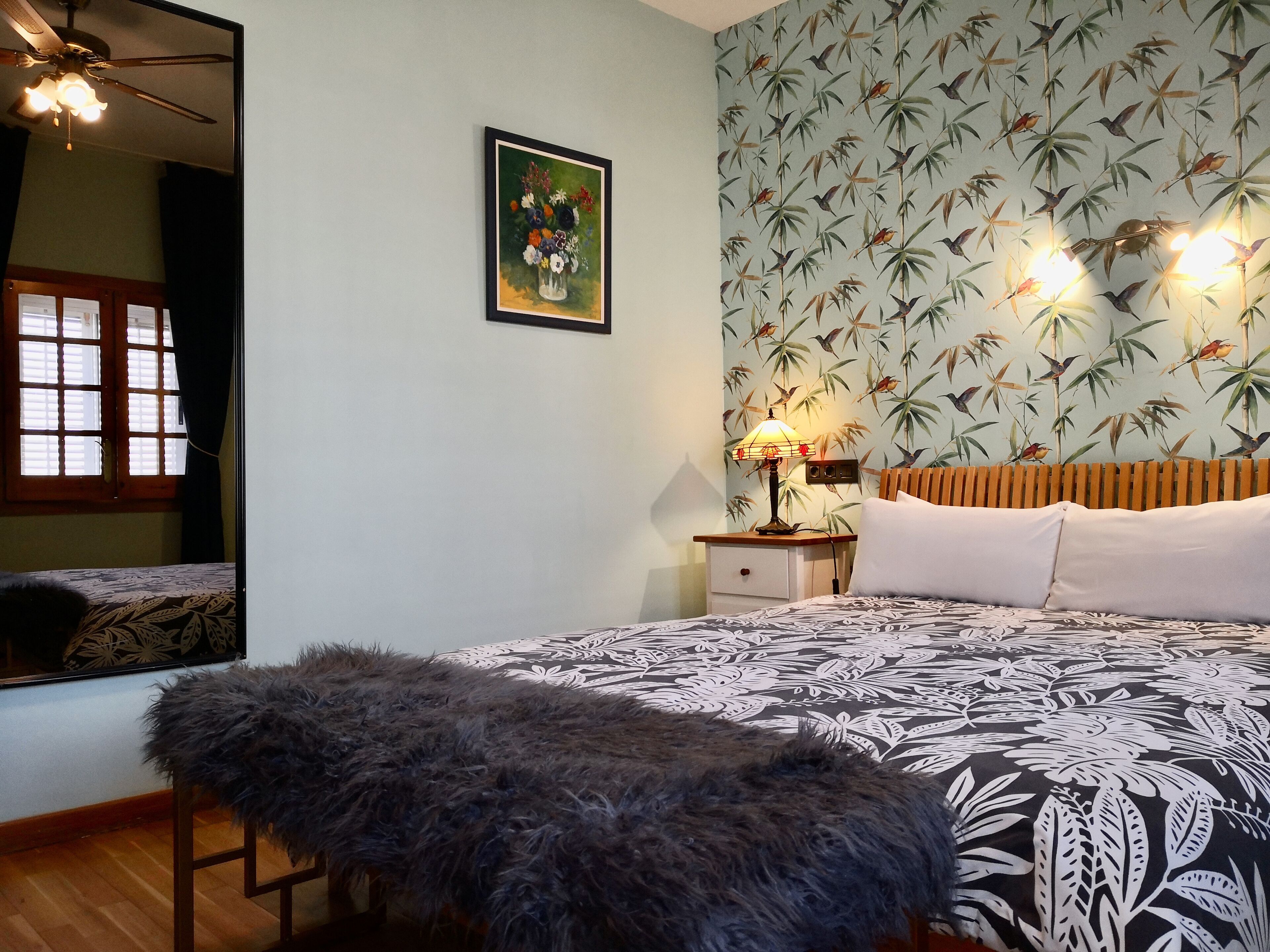 A bedroom with a bed, a nightstand, a lamp, a mirror, and a wall with a bamboo and bird pattern.