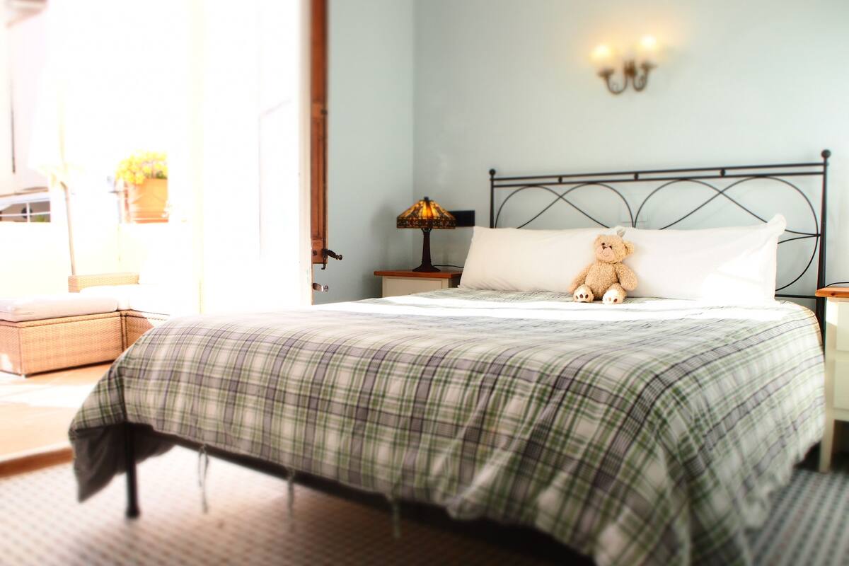 Una cama bien tendida con una colcha escocesa, una lámpara de noche y un osito de peluche sobre la almohada.