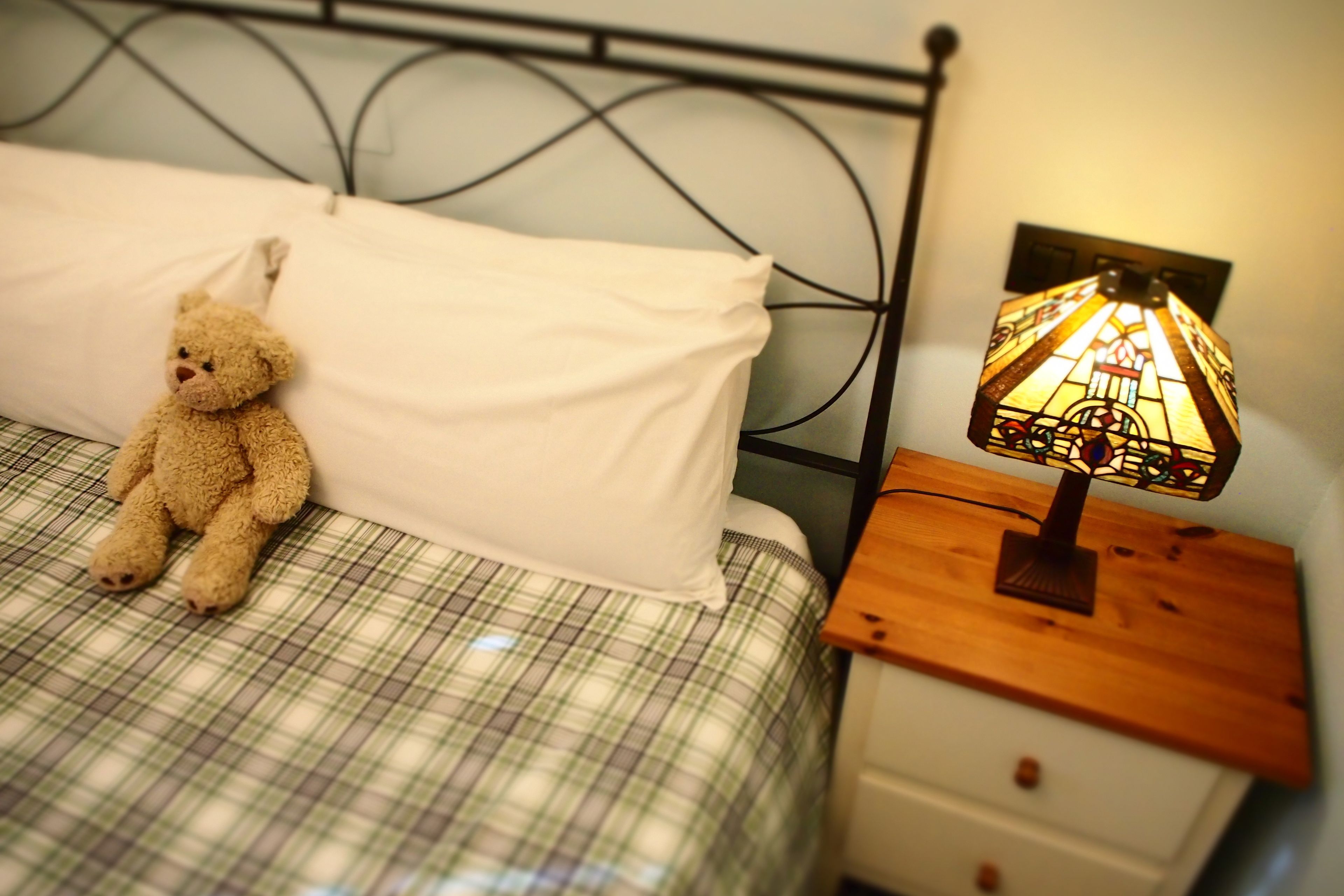 Una cama con una colcha escocesa, un almohilar blanco y un oso de peluche a la izquierda. Junto a la cama, una mesita de noche con una lámpara de vitral.