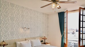 Un dormitorio con cama, mesitas de noche, ventilador de techo, ventana con cortinas y vistas al exterior.