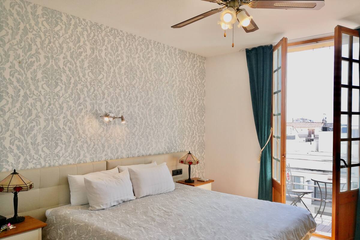 Un dormitorio con cama, mesitas de noche, ventilador de techo, ventana con cortinas y vistas al exterior.