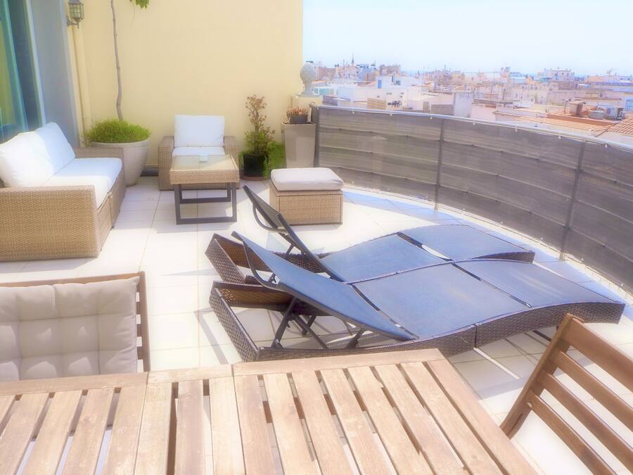 Una terraza en la azotea con mobiliario de mimbre, una tumbona y vistas de la ciudad.