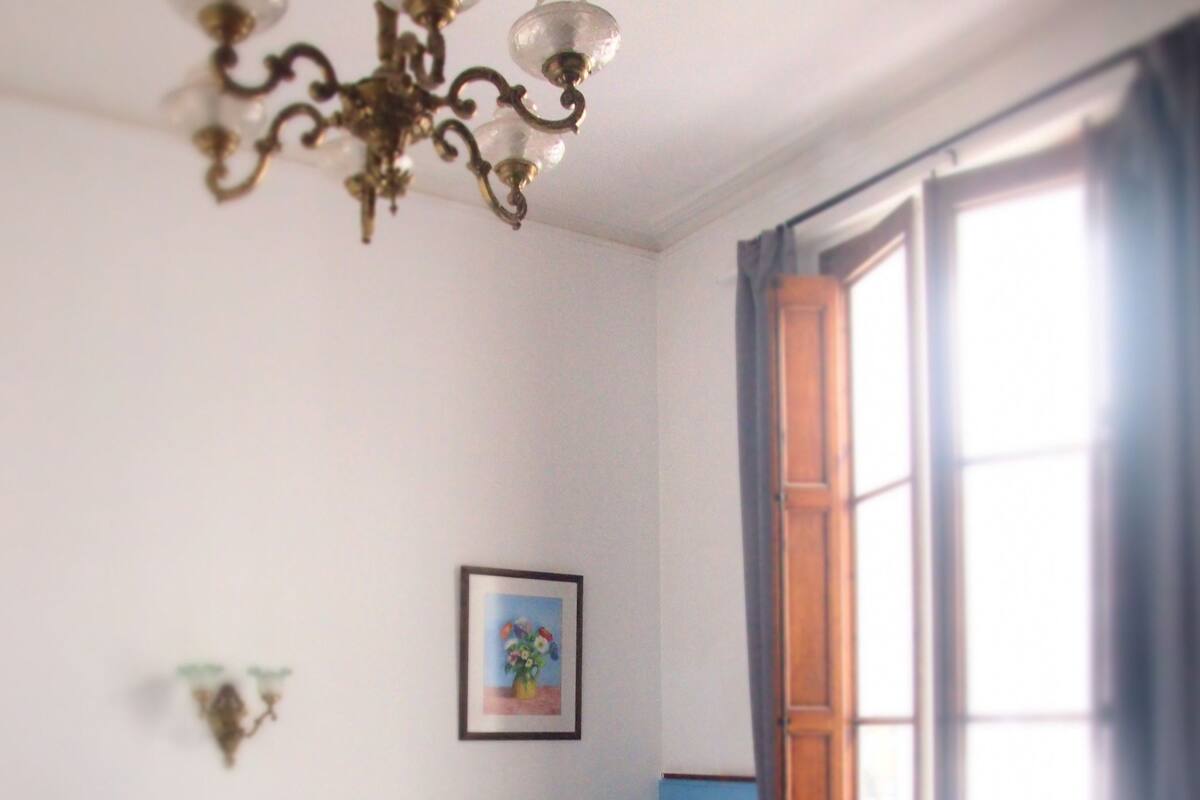 Un dormitorio con cama, una silla, ventana con cortinas, una araña y un cuadro en la pared.