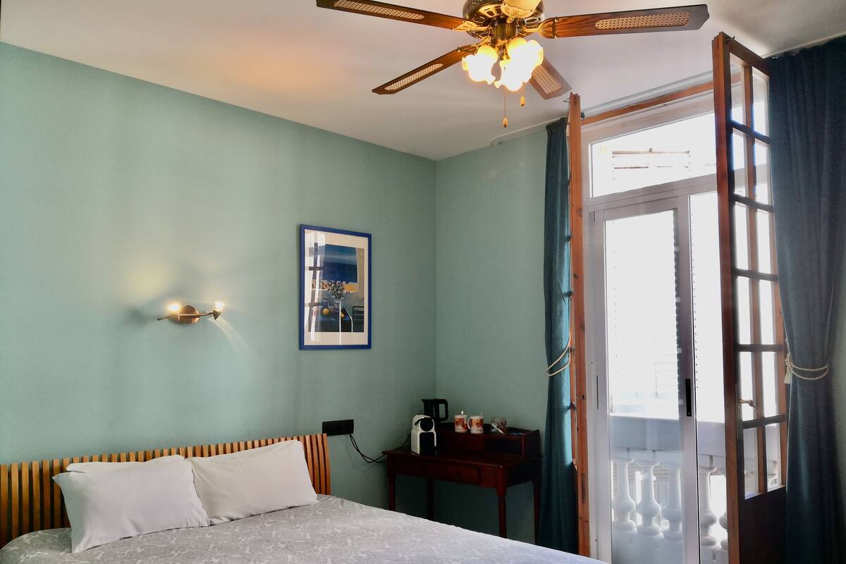 Un dormitorio con cama, ventilador de techo, ventana con cortinas y una mesita de noche con lámpara.