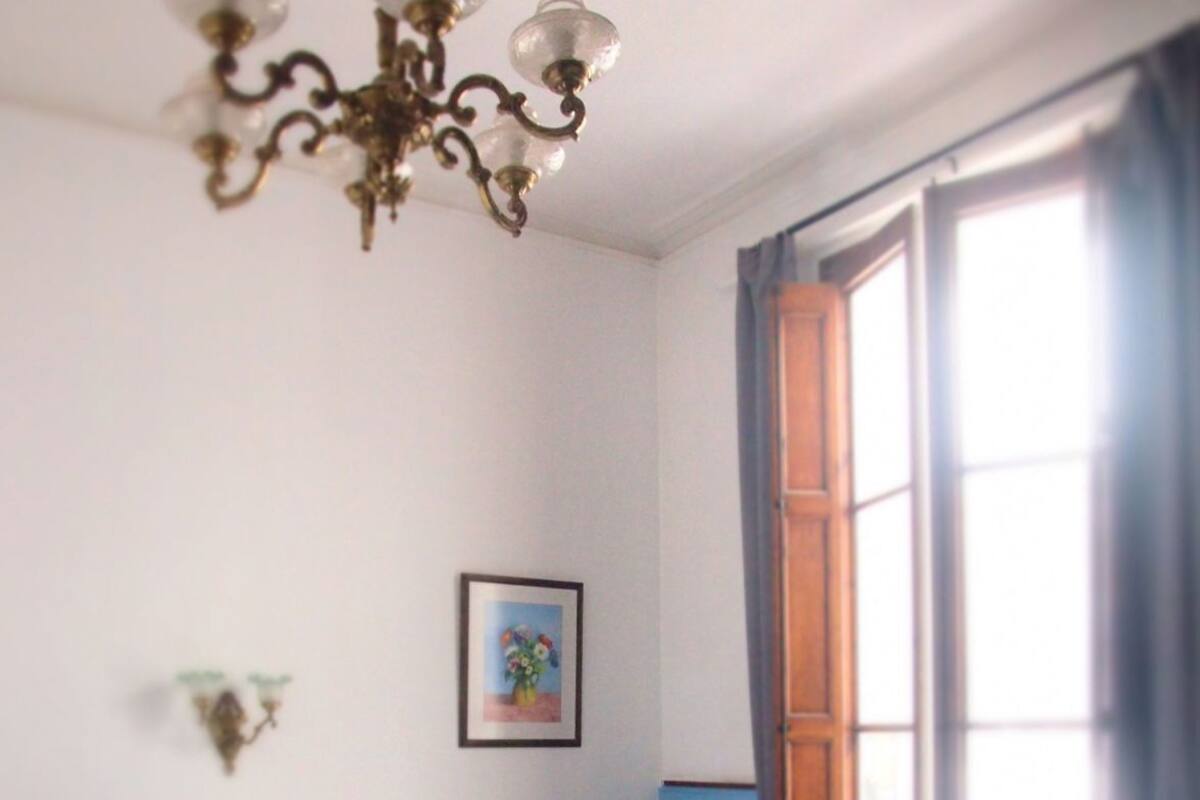 Un dormitorio con cama, una silla, un ventanal y una araña.