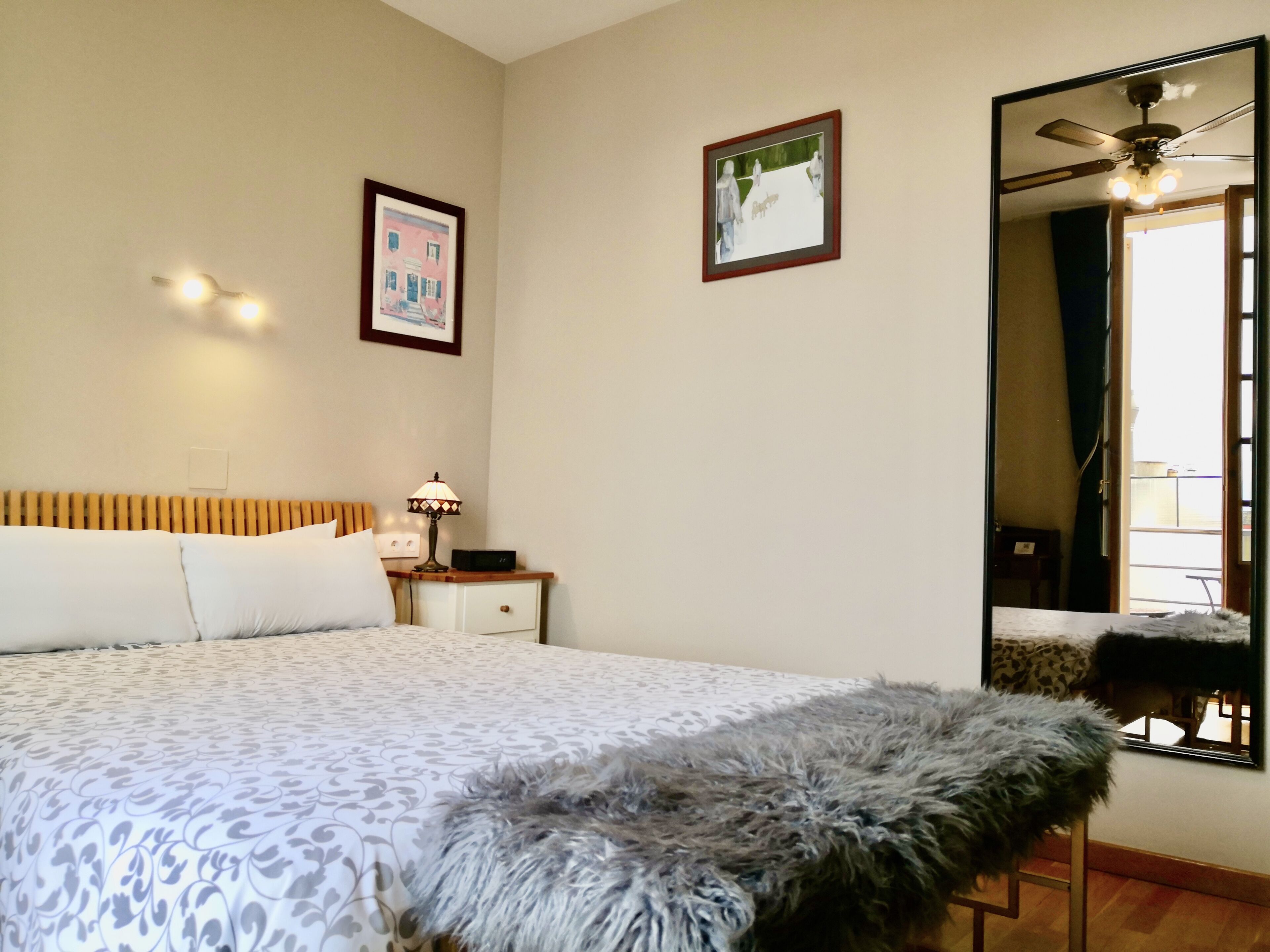 Un dormitorio con cama, mesita de noche, lámpara, un cuadro enmarcado, ventilador de techo y un espejo grande.