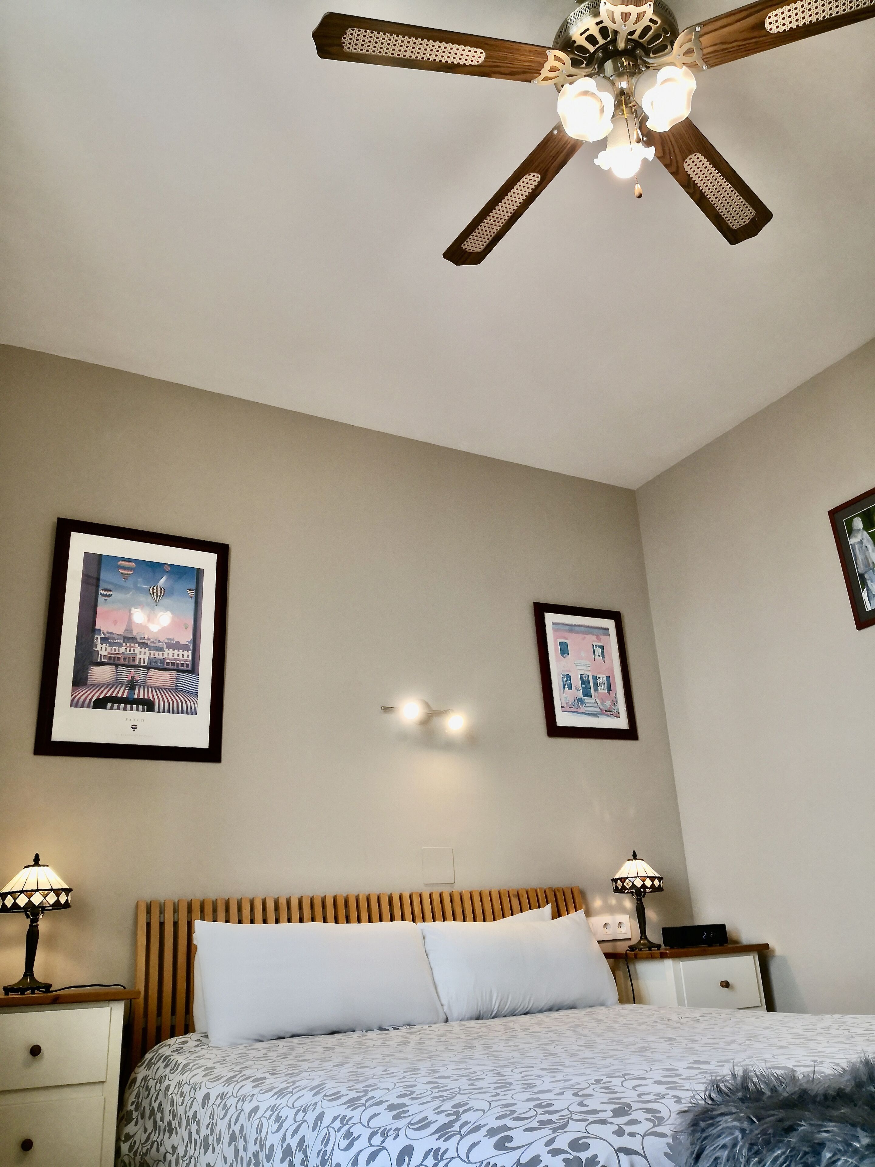 A bedroom with a bed, bedside lamps, a ceiling fan, and framed pictures on the walls.