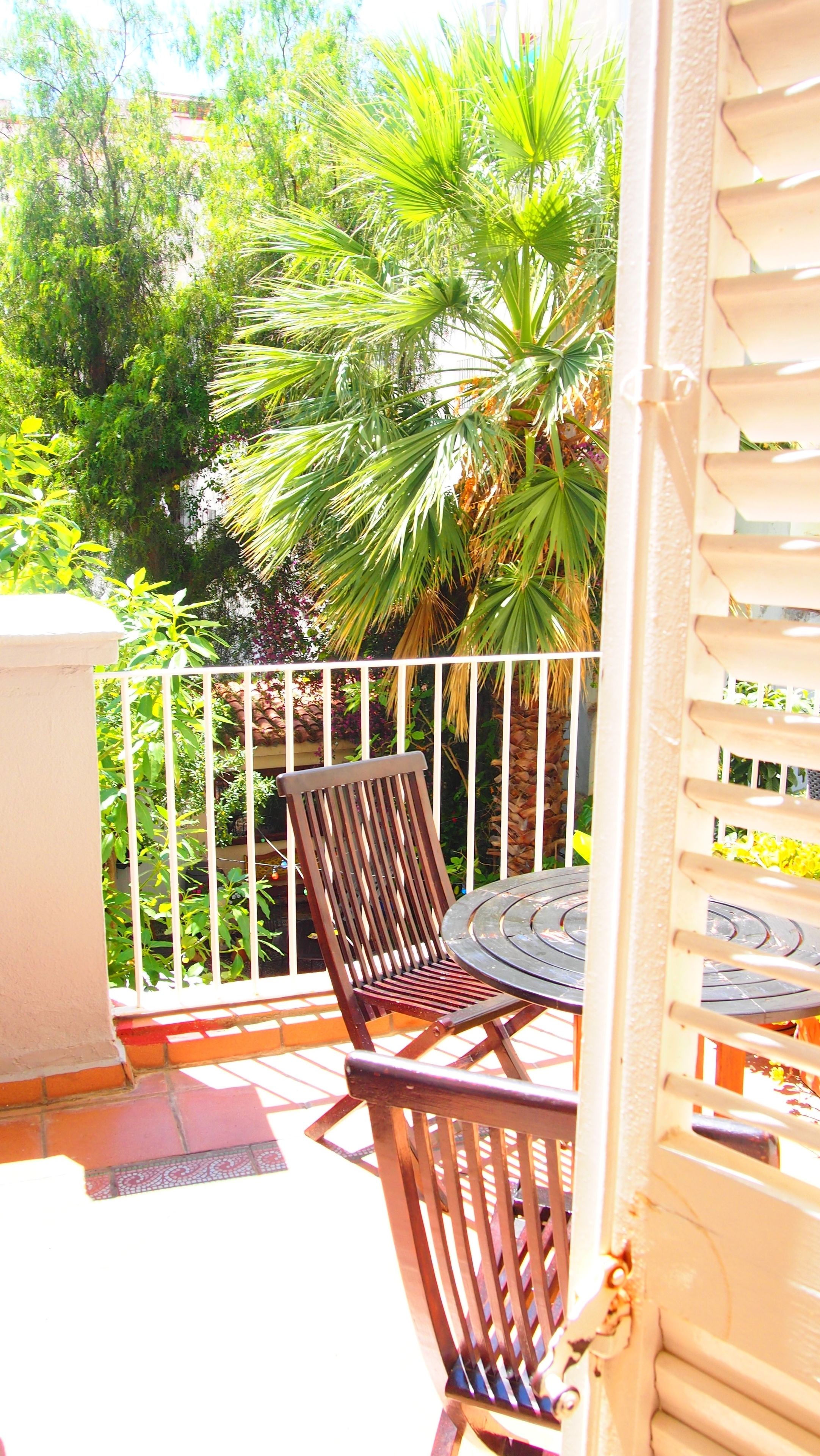 A balcony with a wooden chair and a small round table, surrounded by a white railing and lush greenery.