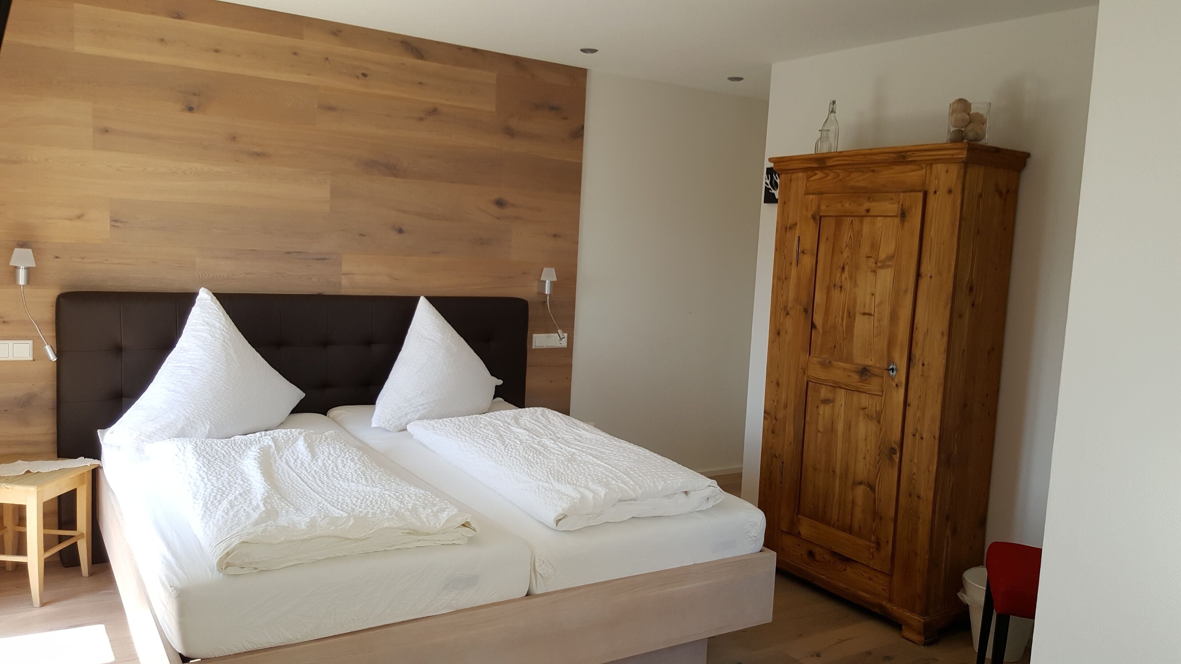 A double bed with white bedding, a wooden headboard, and two bedside lamps. A wooden wardrobe is to the right of the bed.