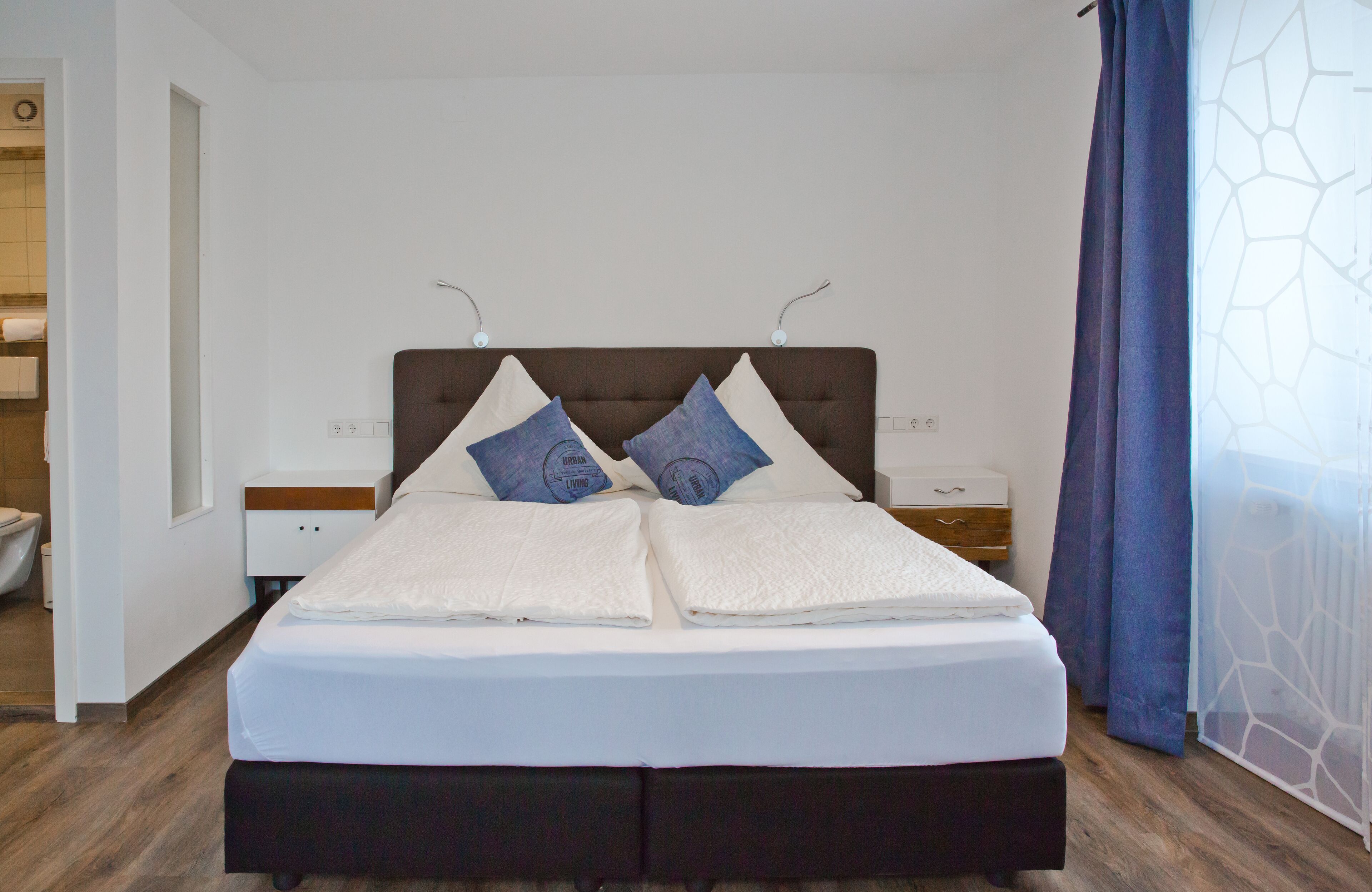 A neatly made bed with white linens and two pillows, a wooden headboard, and a nightstand with a drawer.