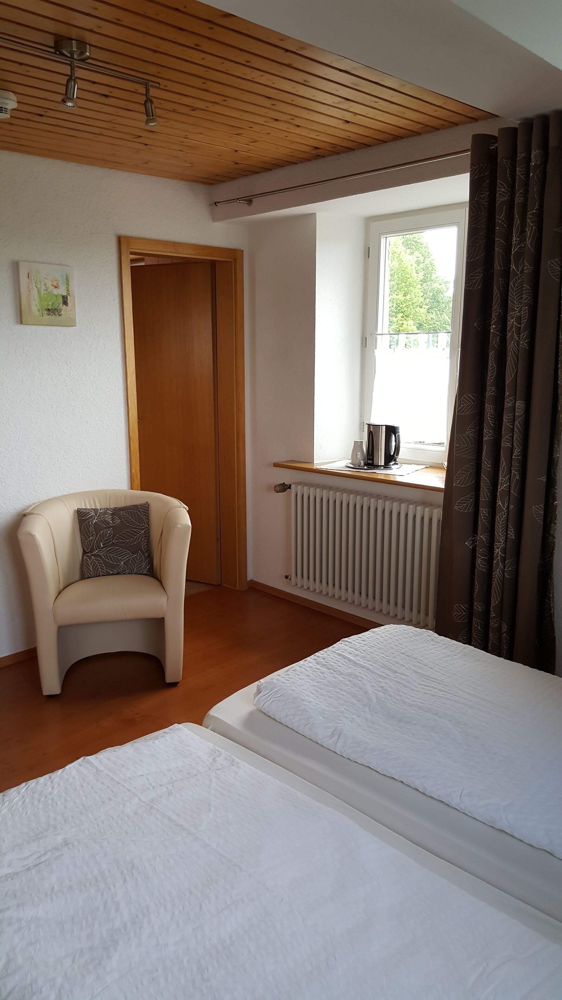 A bedroom with two beds, a wooden ceiling, a radiator, a chair, and a window with curtains.