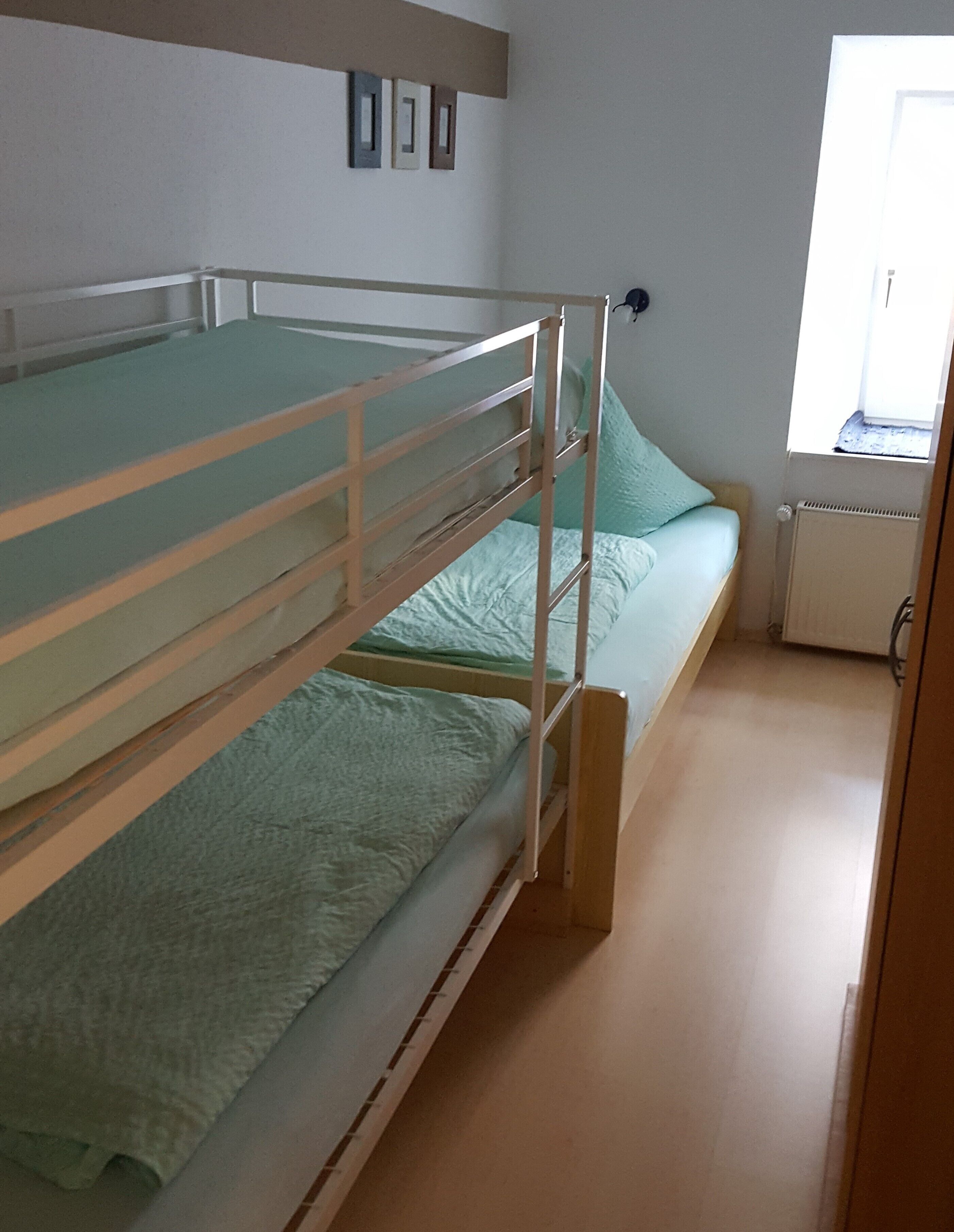 A bunk bed with a green mattress and a single bed with a light blue mattress. There is a window with a view of the outside.