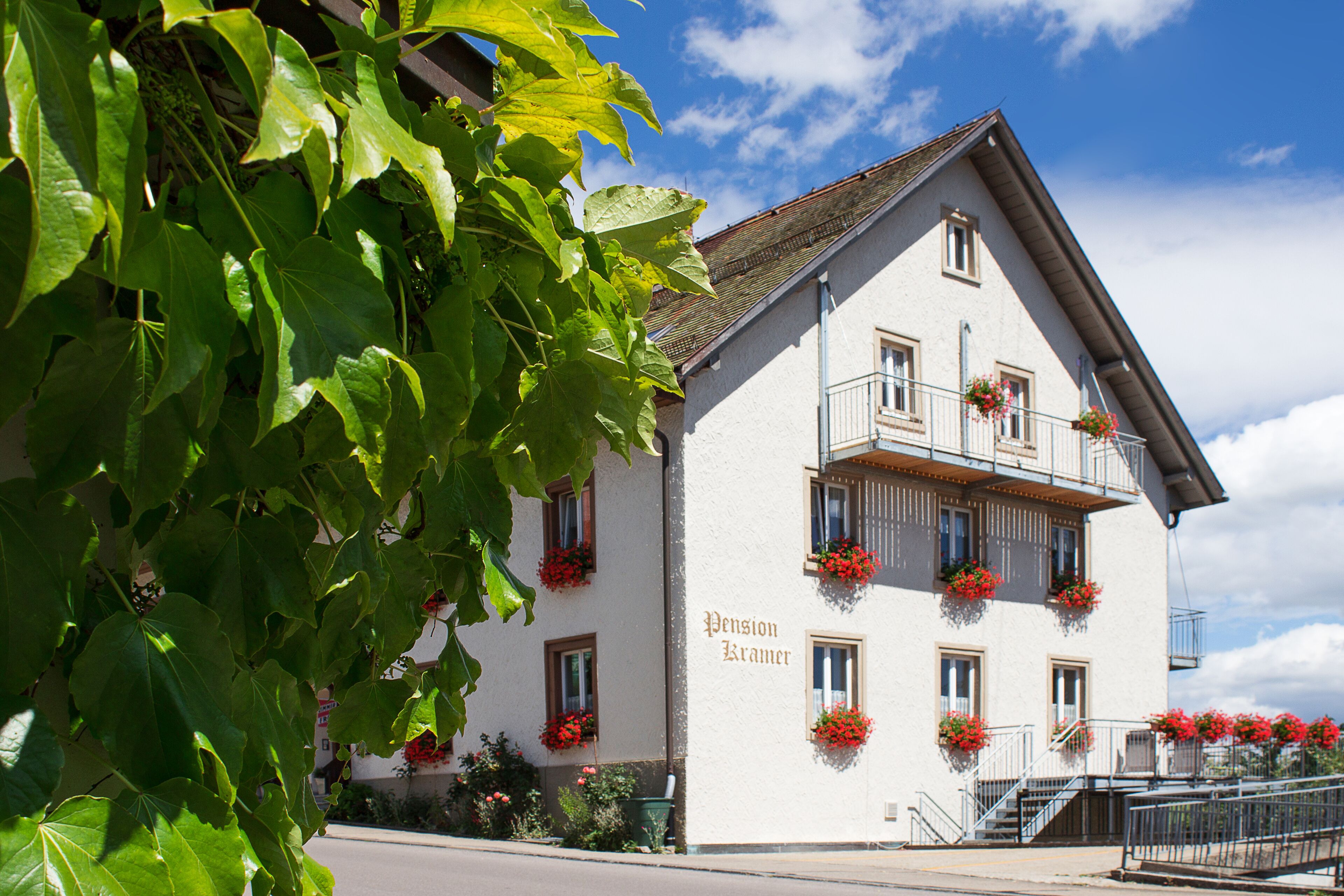 A white building with the name "Pension Kramer" on its facade, featuring multiple windows with flower boxes and a balcony with railings.