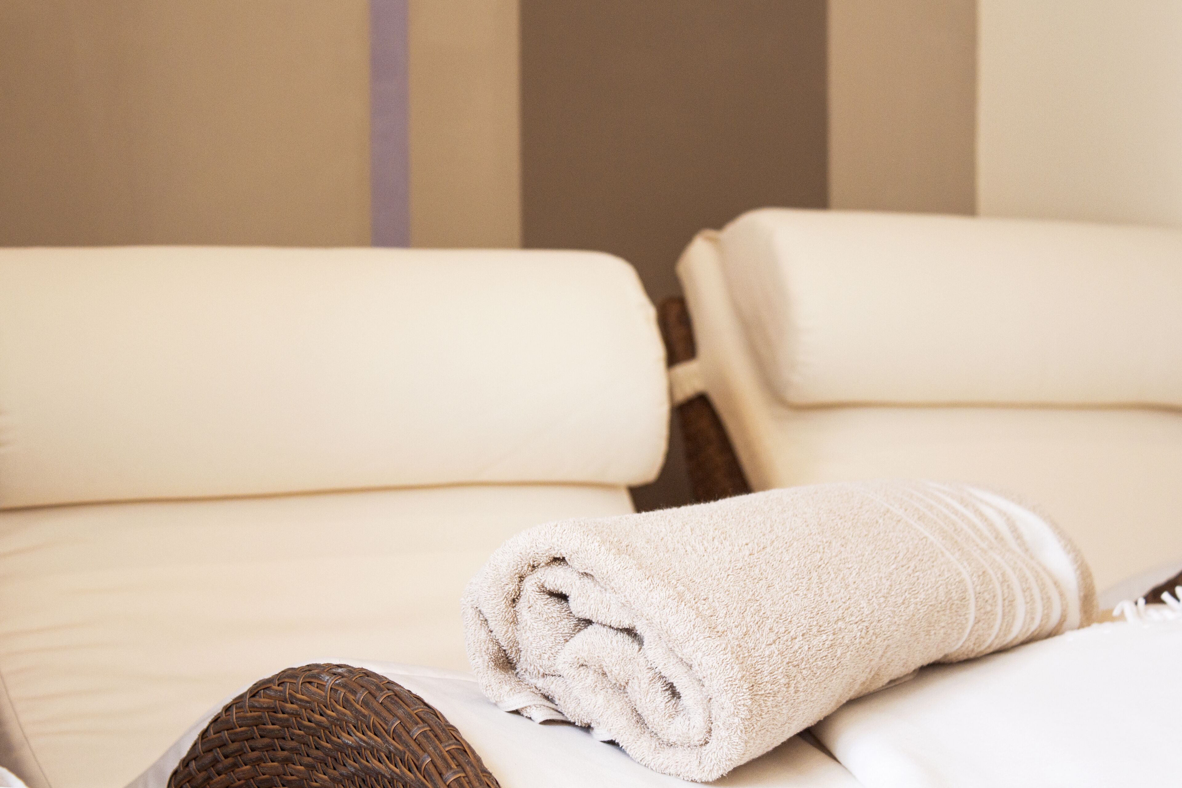 A neatly made bed with a rolled white towel and a woven brown item, likely a blanket or cushion, in a room with neutral-toned walls.