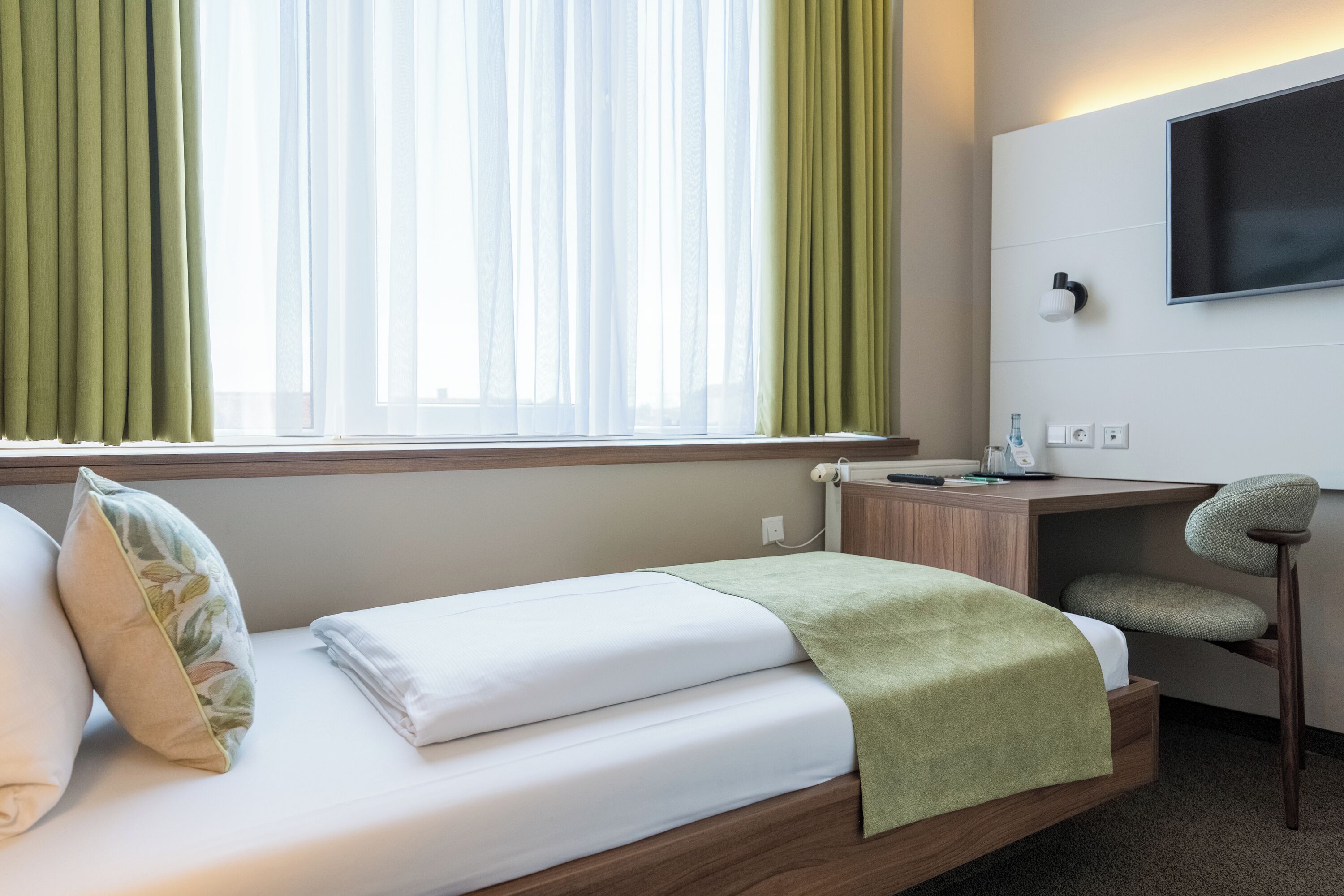 Habitación de hotel con cama, escritorio, silla, televisor montado en la pared y ventana con cortinas.