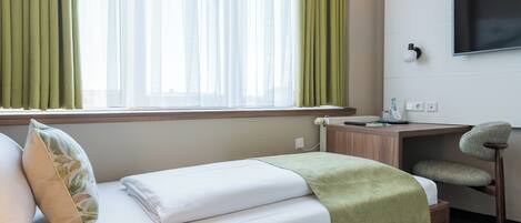 Habitación de hotel con cama, escritorio, silla, televisor montado en la pared y ventana con cortinas.