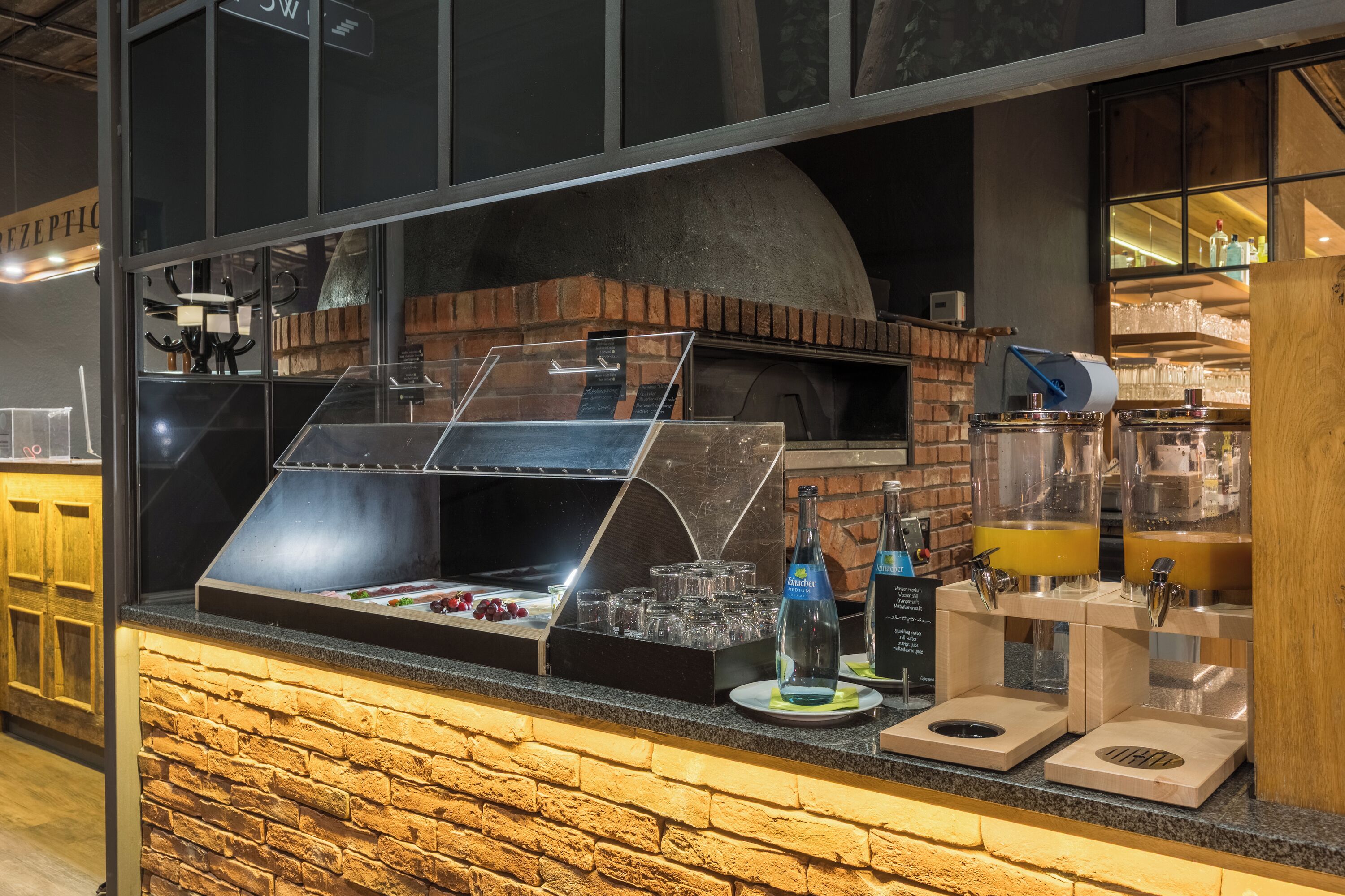 Um balcão de tijolos com uma vitrine de vidro, um forno a lenha e um dispensador de bebidas.