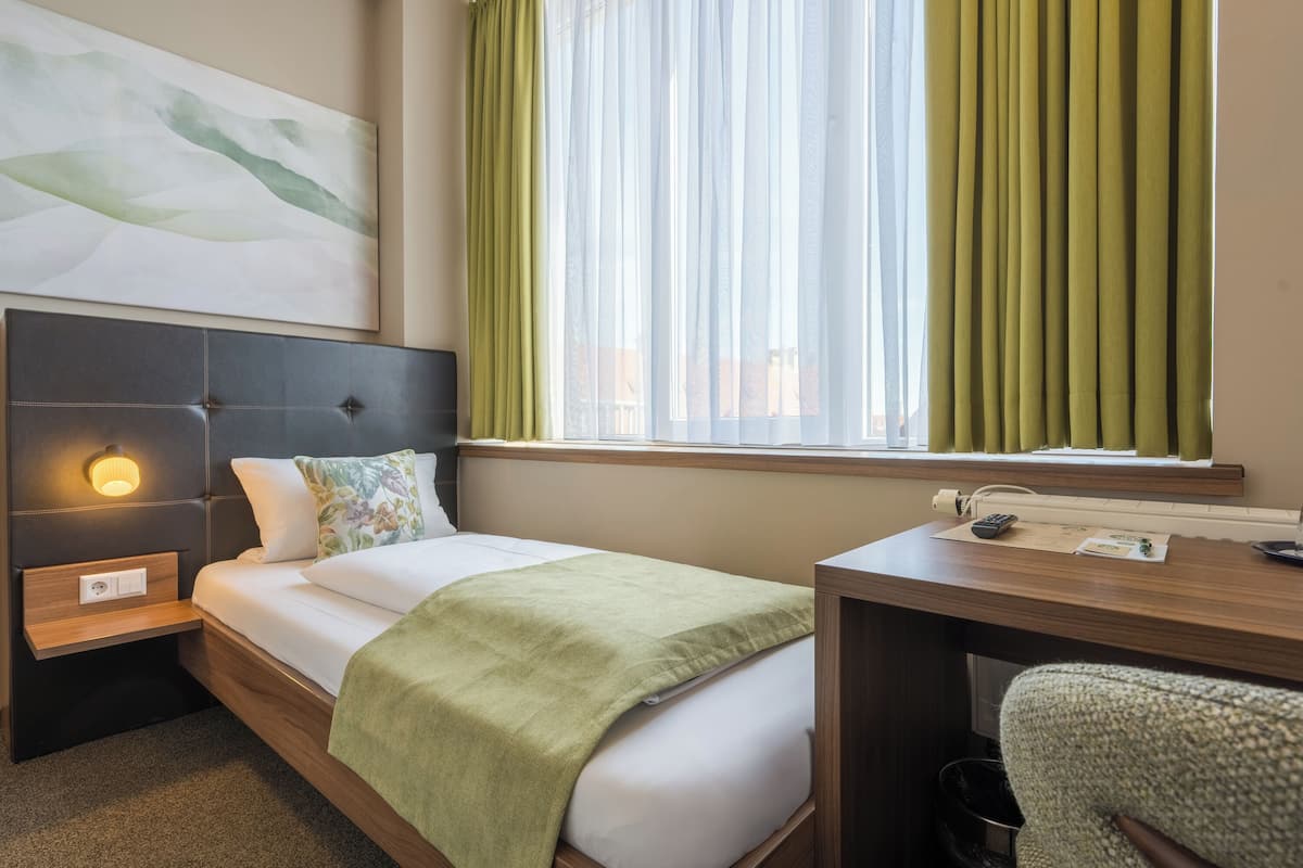 Habitación de hotel con cama, escritorio, silla, mesita de noche, lámpara de pared y ventana con cortinas.