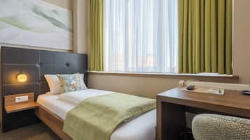 A hotel room with a bed, a desk, a chair, a nightstand, a wall-mounted lamp, and a window with curtains.