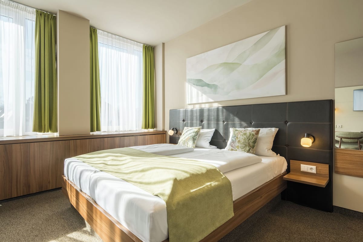 Una habitación de hotel moderna con una cama grande, una mesita de noche con lámpara, una ventana con cortinas y una obra de arte colgada en la pared.
