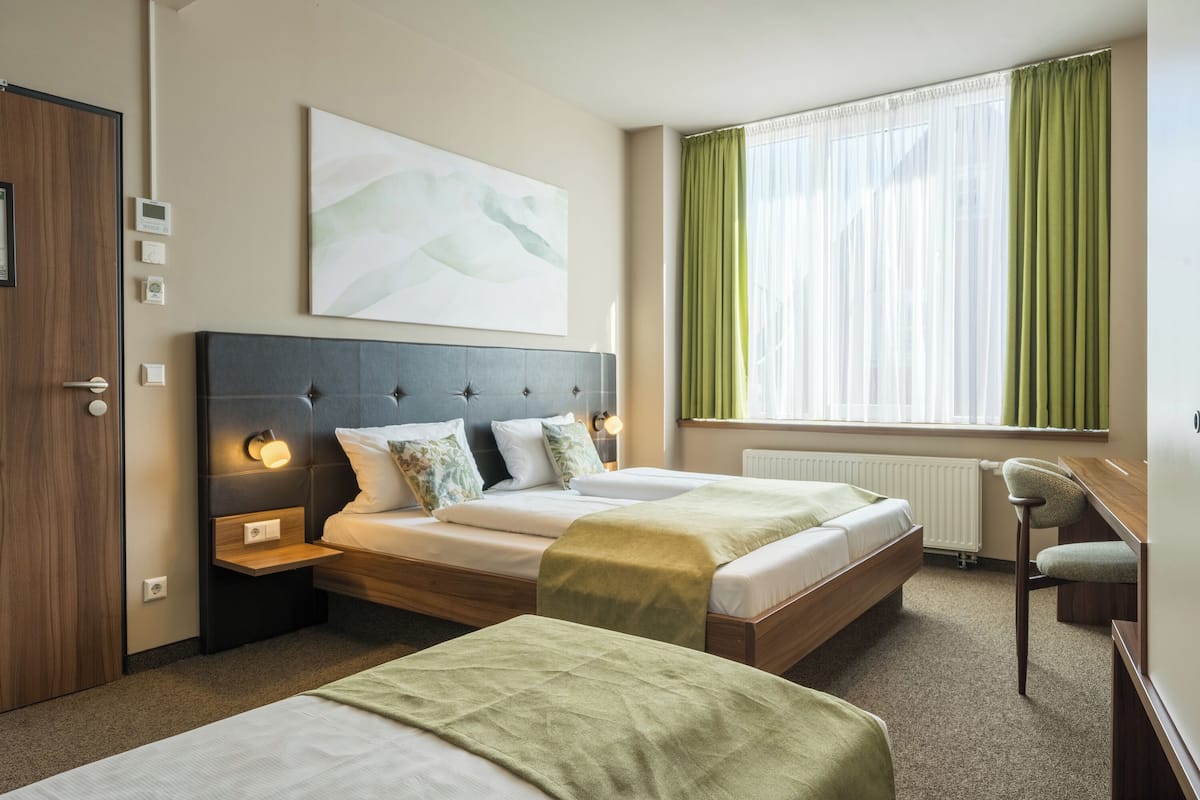 Habitación de hotel con dos camas, un escritorio, una silla, una ventana con cortinas y un cuadro en la pared.