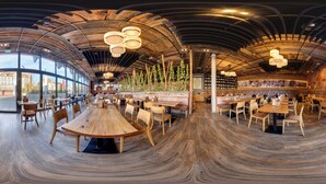 Un restaurante espacioso con mesas y sillas de madera, una pared de ladrillo con plantas y un techo curvo con luces colgantes.