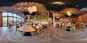 A spacious restaurant with wooden tables and chairs, a brick wall with greenery, and a curved ceiling with hanging lights.