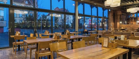 Un restaurante con mesas y sillas de madera, grandes ventanales y vistas a edificios.