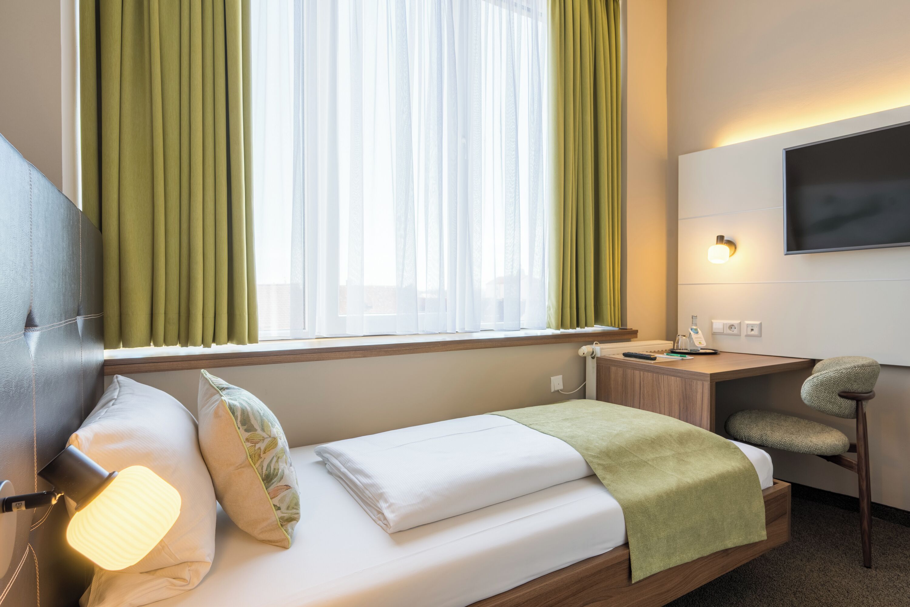A hotel room with a bed, bedside lamp, green curtains, a desk with a chair, and a TV mounted on the wall.