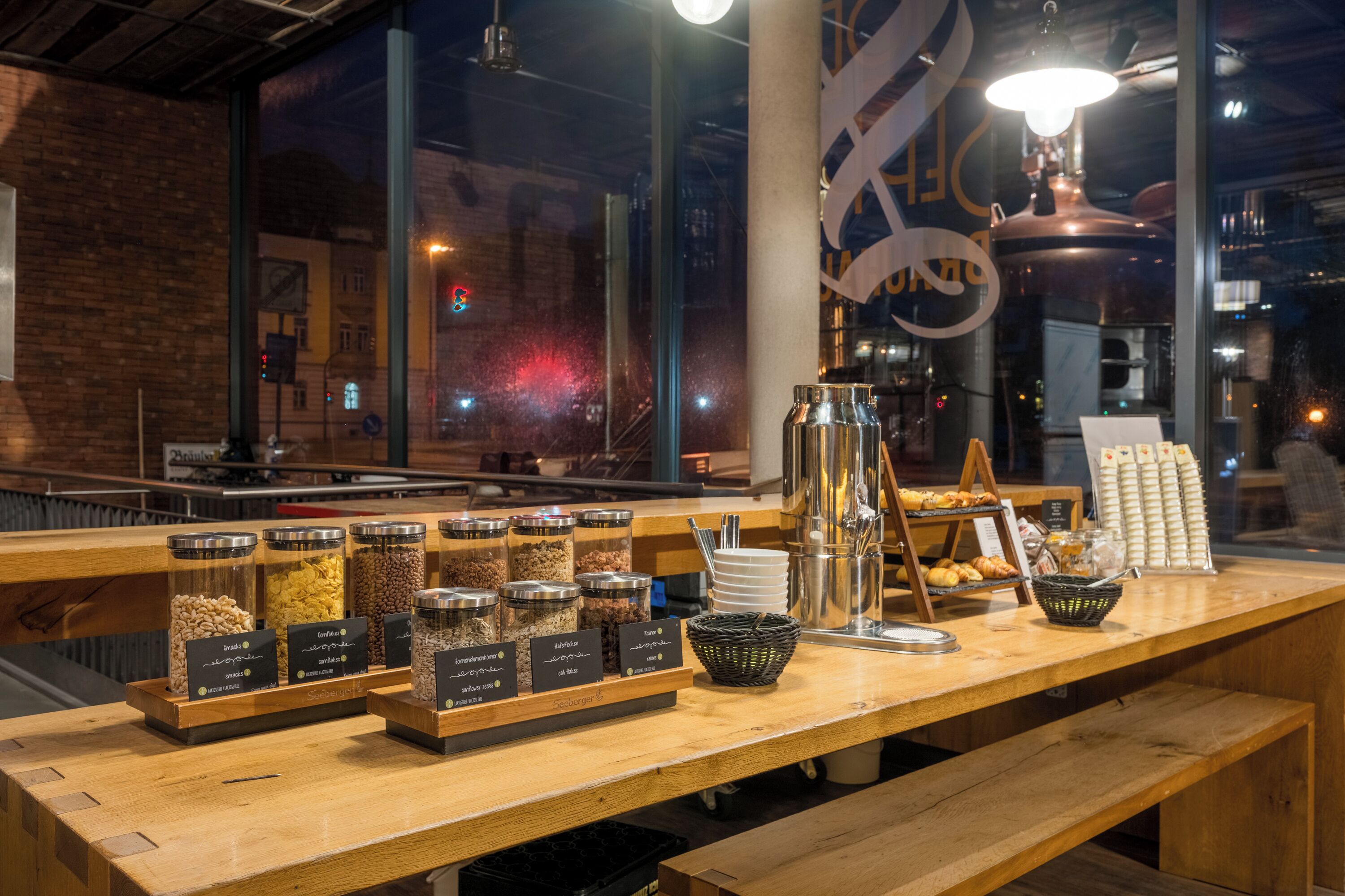 Ein Frühstücksbuffet mit Gläsern voller Granola und anderen Frühstücksartikeln, eine Kaffeemaschine und Blick auf die Stadt durch große Fenster.