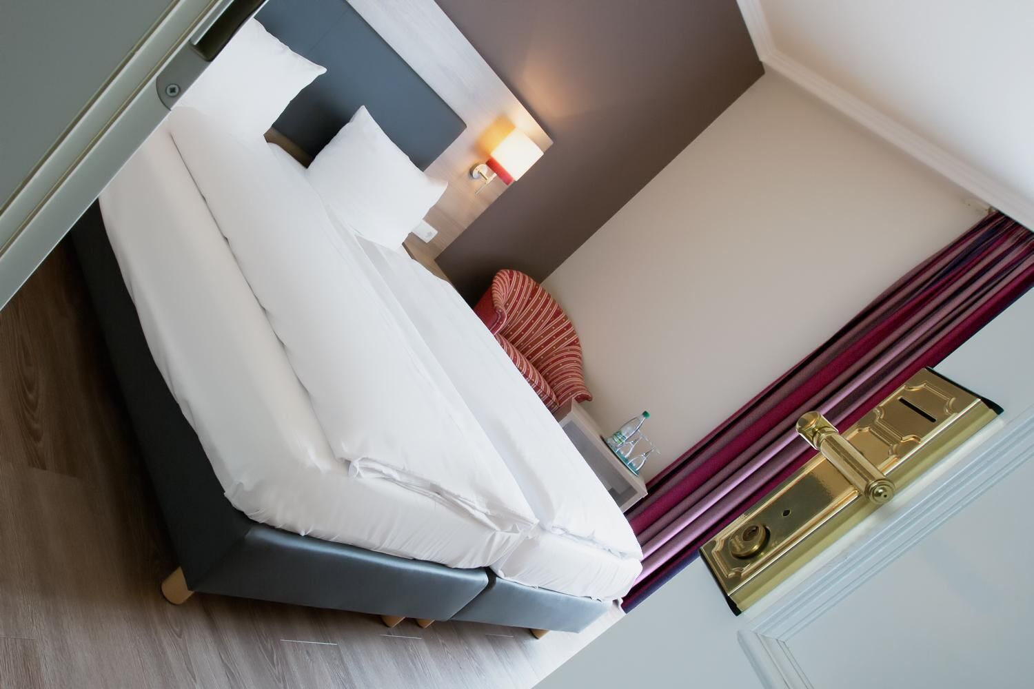 Habitación de hotel con cama, mesita de noche, lámpara, botella de agua y una puerta con pomo dorado.