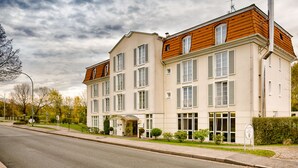 Un hôtel de deux étages, de style architectural européen classique, caractérisé par une façade symétrique, de nombreuses fenêtres avec volets et une entrée centrale surmontée d’un auvent.