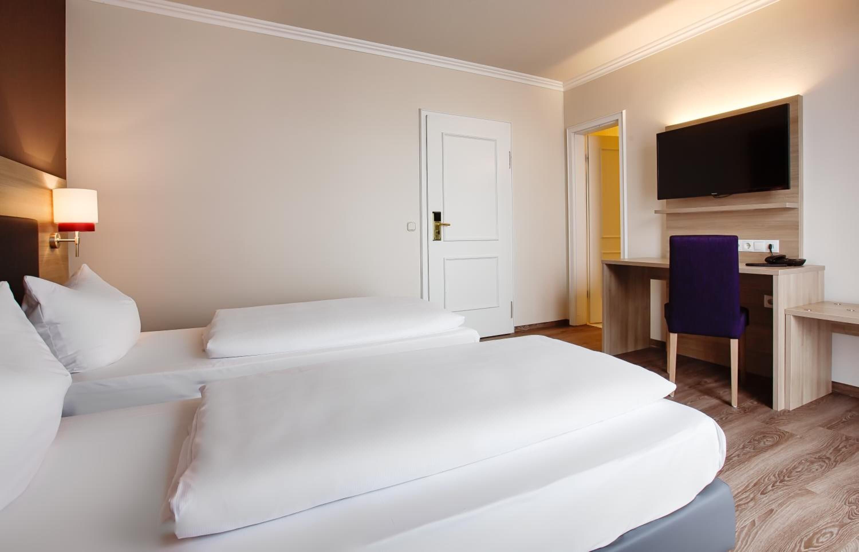 A hotel room with two beds, a desk, a chair, a TV, and a door.