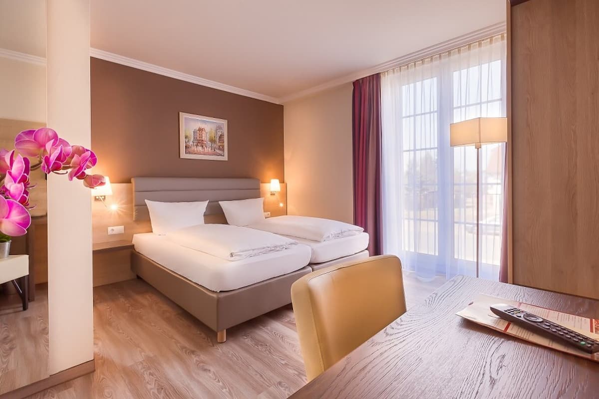 Ein Hotelzimmer mit einem Bett, Nachttischlampen, einem Tisch mit Fernbedienung und einem Fenster mit Vorhängen.