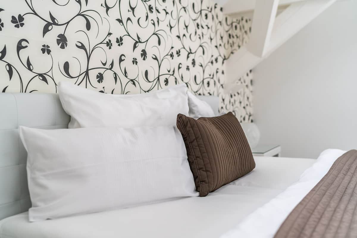 A neatly made bed with white and brown pillows, a white headboard, and a patterned wallpaper.
