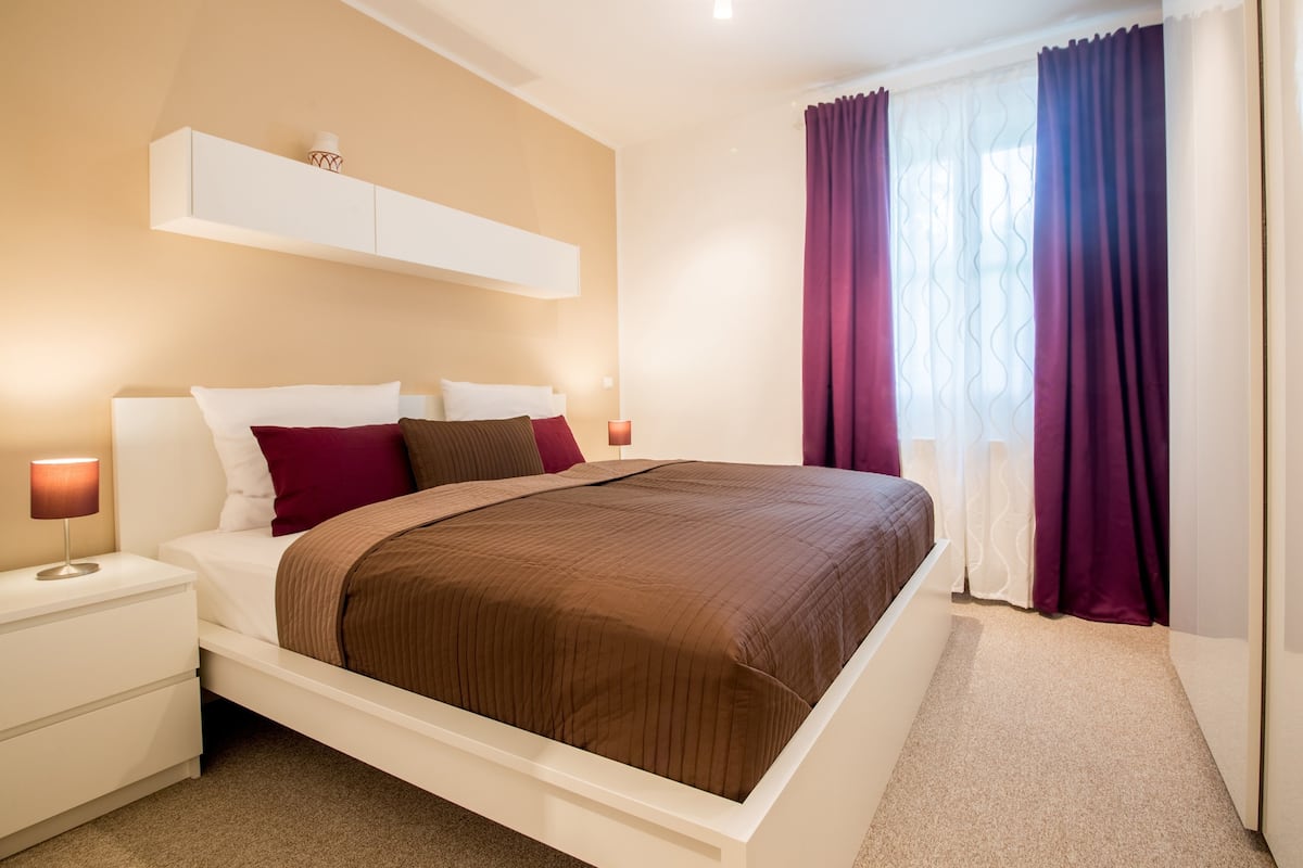 A modern bedroom with a large bed, white nightstands, a bedside table with a lamp, and a window with purple curtains.