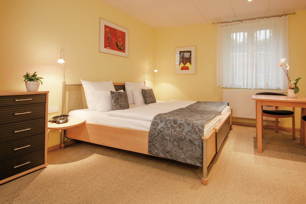 A neatly arranged bedroom with a bed, bedside table, lamp, dresser, and a window with curtains.