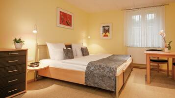 A neatly arranged bedroom with a bed, bedside table, lamp, dresser, and a window with curtains.
