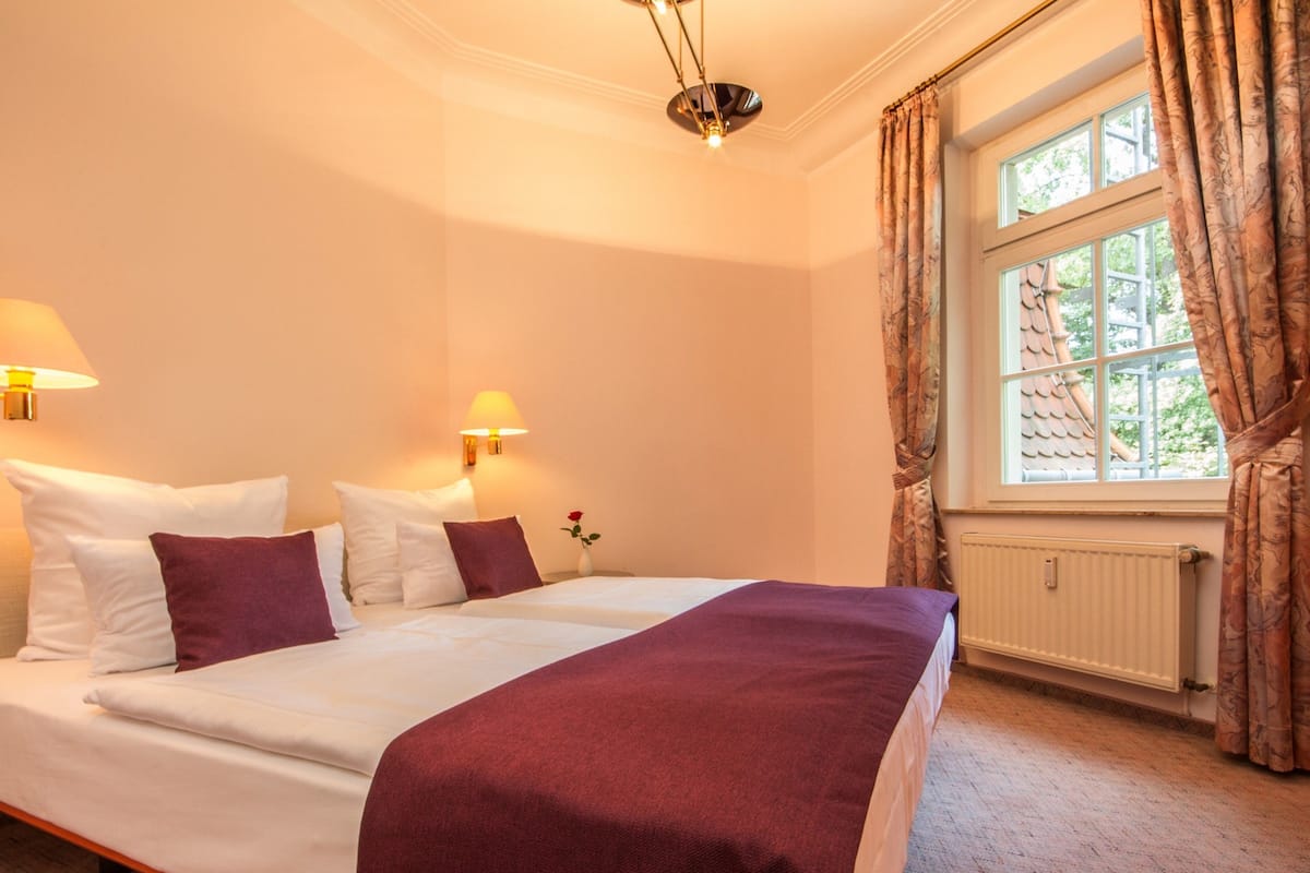 A hotel room with a large bed, two bedside lamps, a window with curtains, and a radiator.