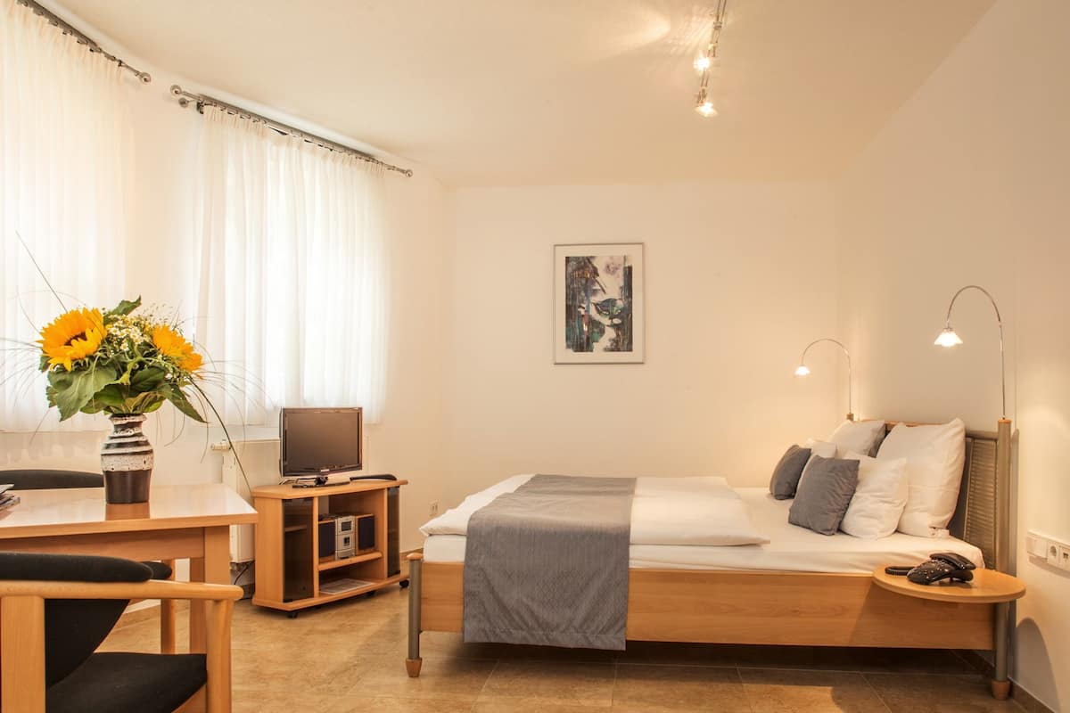 A hotel room with a bed, bedside table, lamp, desk, chair, TV, and a vase with flowers.
