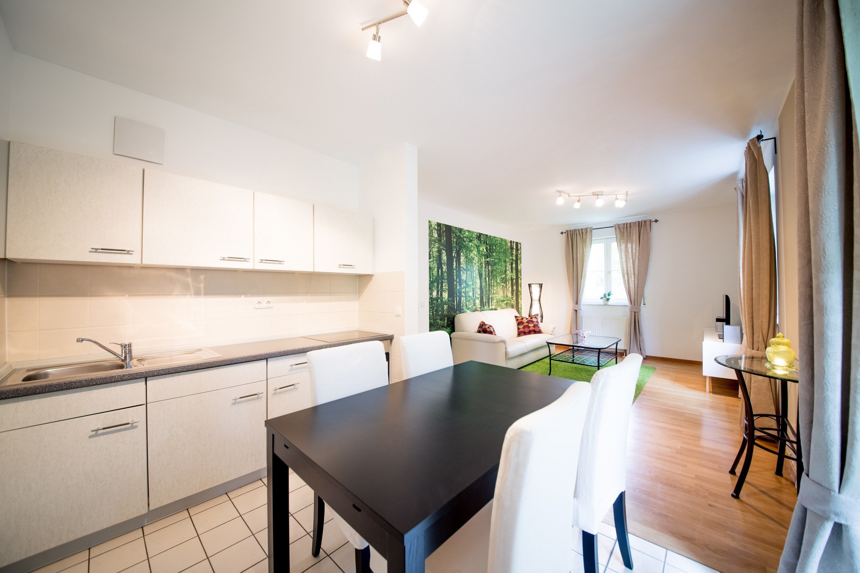 A modern kitchen with white cabinets, a black dining table, and a living area with a sofa and a green wall.