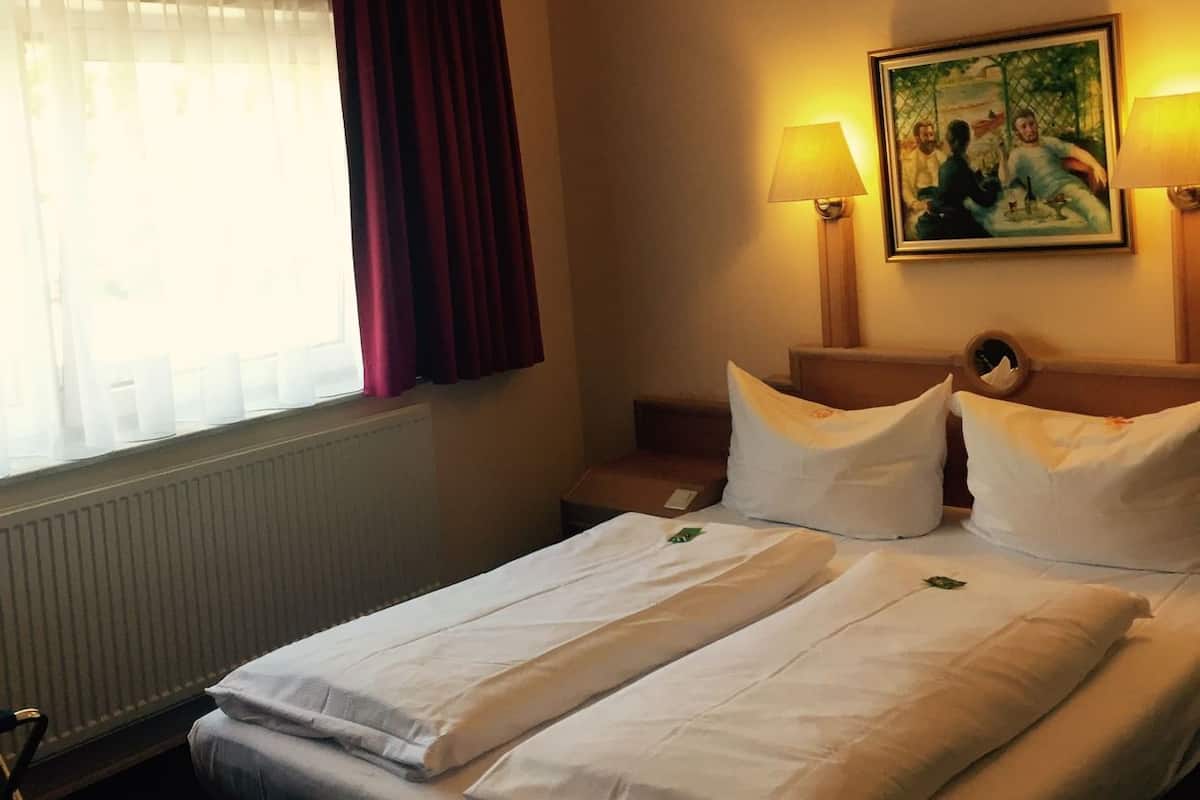 A hotel room with a bed, two bedside lamps, a painting on the wall, and a window with curtains.