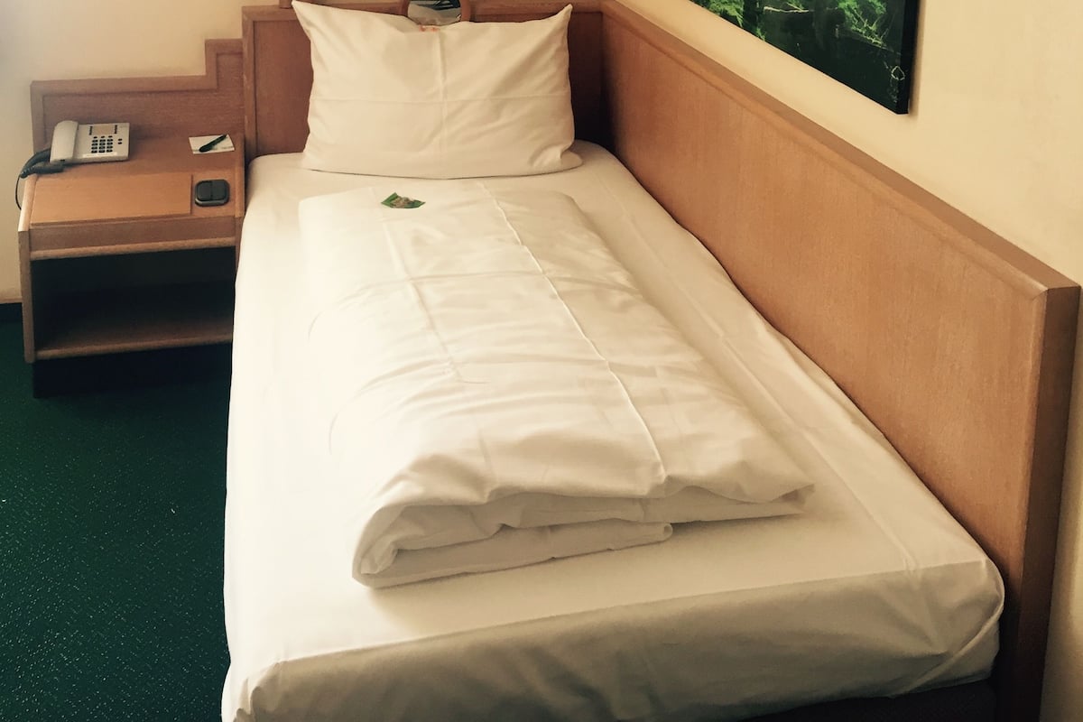 A hotel room with a neatly made bed, a bedside lamp, a phone on the nightstand, and a painting of a forest on the wall.