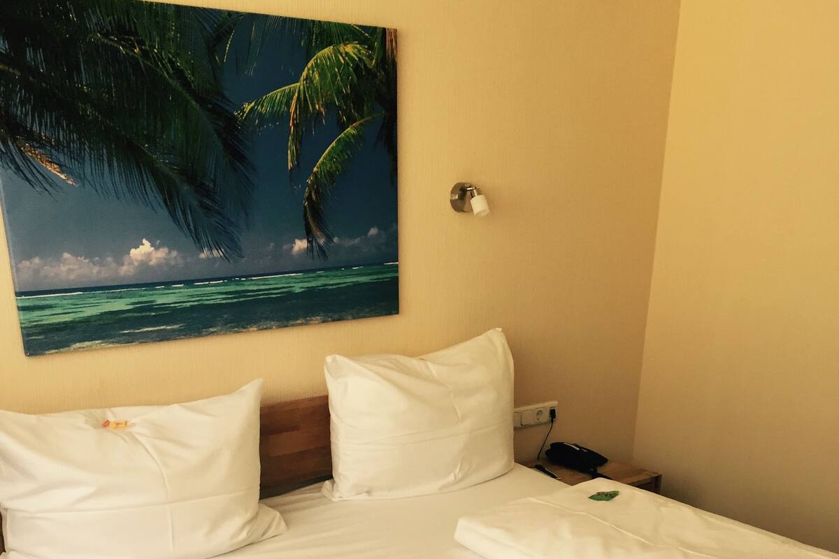 A hotel room with a bed, white linens, a painting of a beach scene, and a nightstand with a lamp.